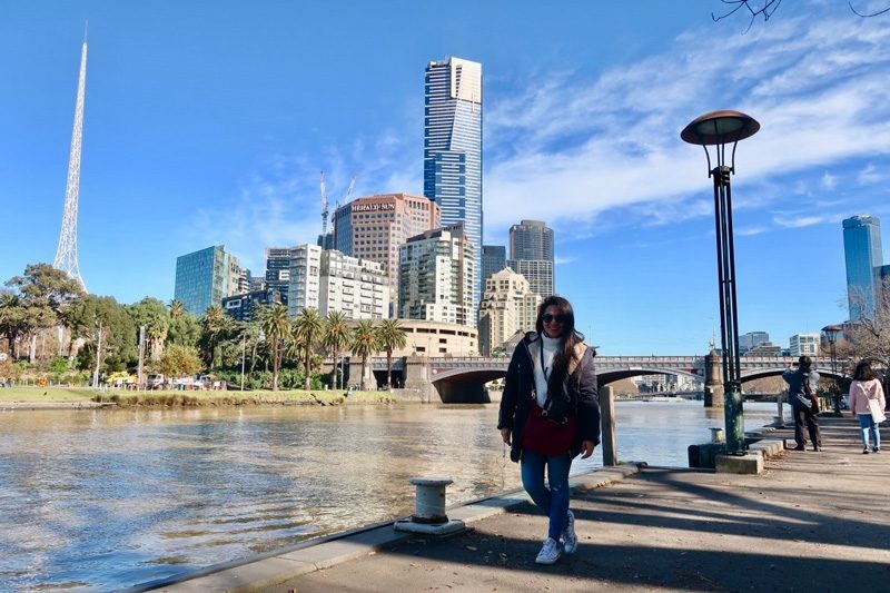 chica morena con abrigo y bambas posando delante de la ciudad de Brisbane en Australia. Detrás de ella hay el río con un puente y los rascacielos de la ciudad